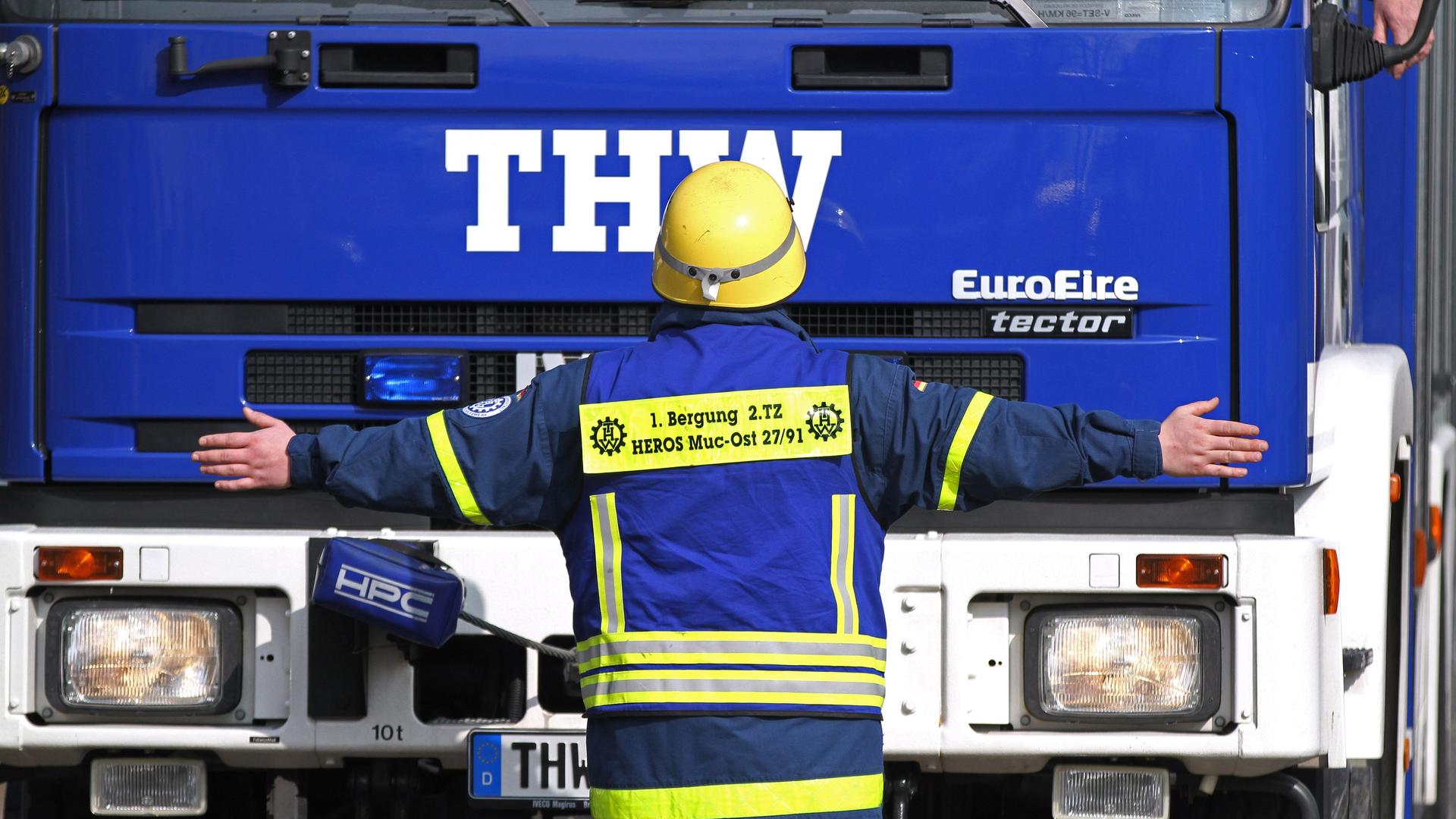 Ein freiwilliger THW-Helfer steht während einer Übung vor einem blauen Lkw des Technischen Hilfswerks. Dabei hat er die Arme ausgestreckt. Ein freiwilliger THW-Helfer steht während einer Übung vor einem blauen Lkw des Technischen Hilfswerks. Dabei hat er die Arme ausgestreckt.