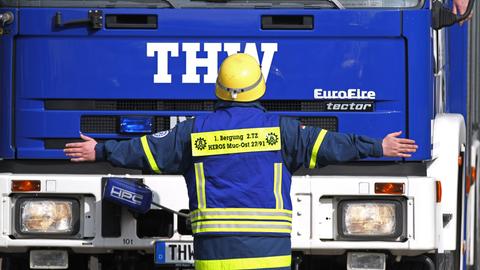 Ein freiwilliger THW-Helfer steht während einer Übung vor einem blauen Lkw des Technischen Hilfswerks. Dabei hat er die Arme ausgestreckt.