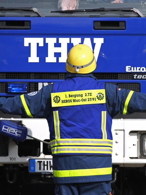 Ein freiwilliger THW-Helfer steht während einer Übung vor einem blauen Lkw des Technischen Hilfswerks. Dabei hat er die Arme ausgestreckt.