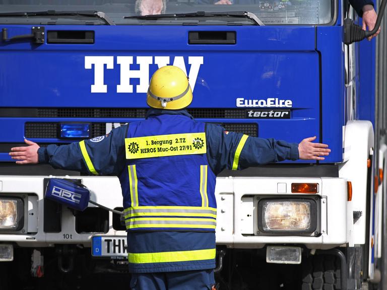Ein freiwilliger THW-Helfer steht während einer Übung vor einem blauen Lkw des Technischen Hilfswerks. Dabei hat er die Arme ausgestreckt.