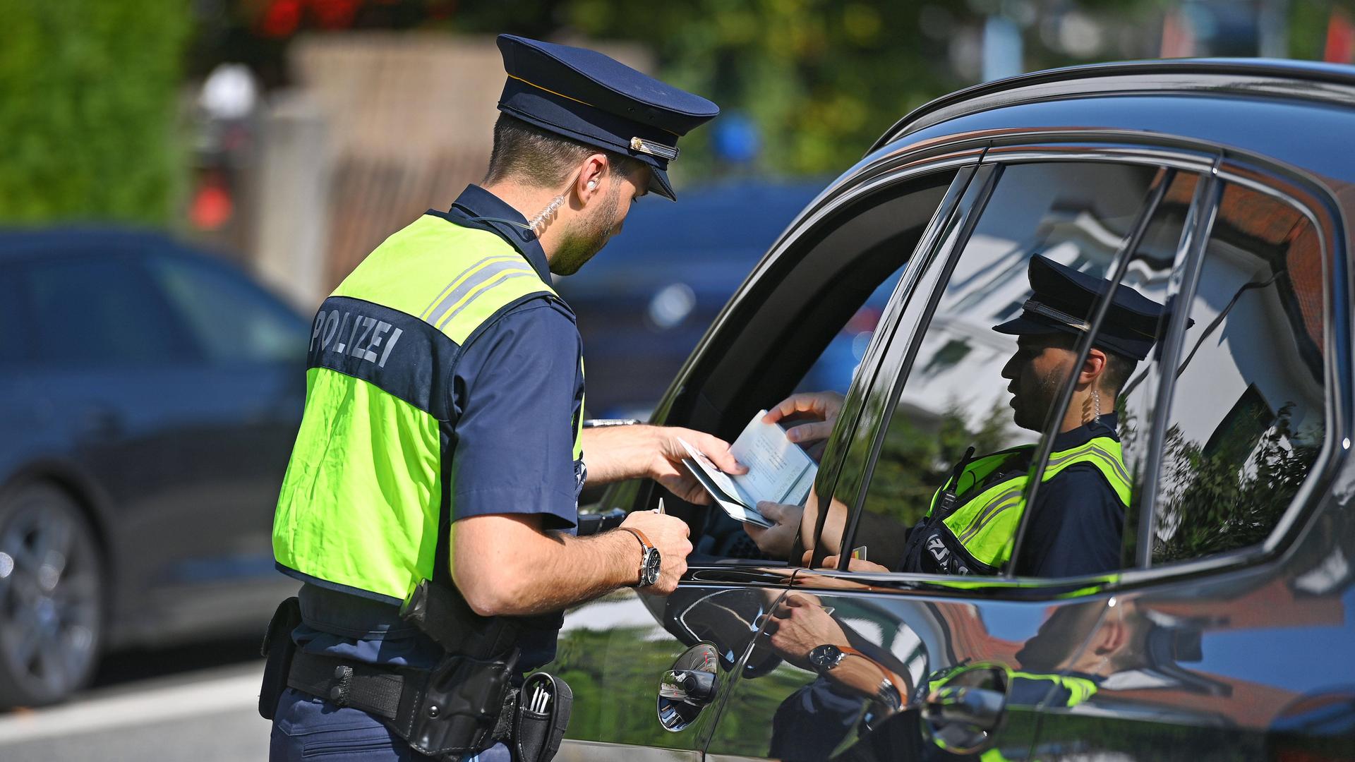 Ein Polizist steht neben einem Auto. Das Fenster an der Fahrer-Tür ist offen. Der Fahrer gibt dem Polizisten einen Pass.