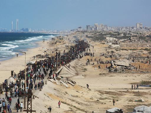 Eine breite Schlange von Menschen läuft einen Standstreifen inmitten des zerstörten Gazastreifens, links das Meer, hinten Ruinen von Gaza-Stadt.