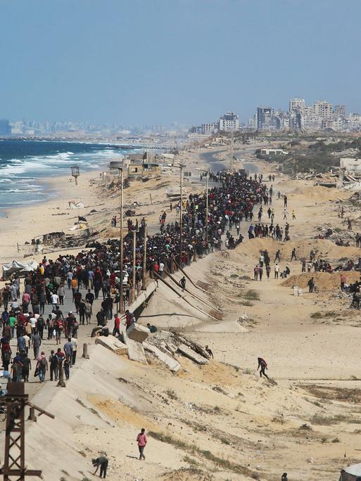 Eine breite Schlange von Menschen läuft einen Standstreifen inmitten des zerstörten Gazastreifens, links das Meer, hinten Ruinen von Gaza-Stadt.