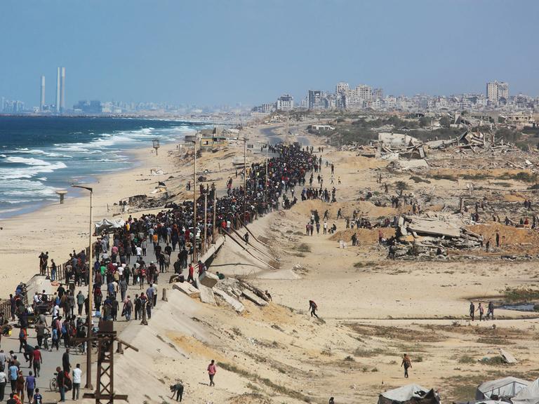Eine breite Schlange von Menschen läuft einen Standstreifen inmitten des zerstörten Gazastreifens, links das Meer, hinten Ruinen von Gaza-Stadt.