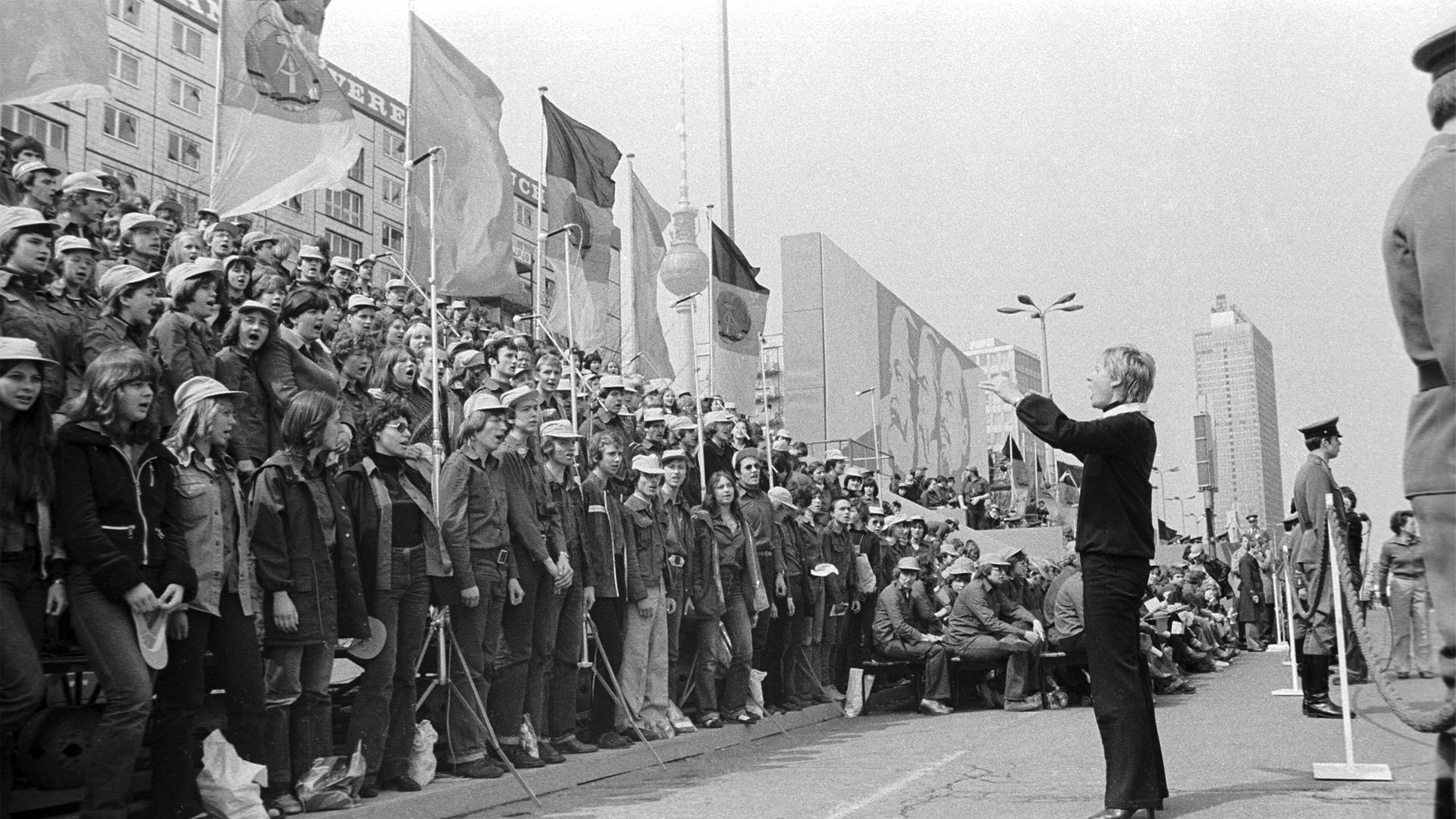 01.05.1980 DDR Ost-Berlin Stadtbezirk Mitte. Szene am Rande der Demonstration zum Ersten Mai. Eine junge Frau dirigiert während des Demonstrationszuges in der Karl-Marx-Allee einen Chor von Angehörigen der Freien Deutschen Jugend. , Berlin 01.05.1980 DDR Ost-Berlin Stadtbezirk Mitte. Szene am Rande der Demonstration zum Ersten Mai. Eine junge Frau dirigiert während des Demonstrationszuges in der Karl-Marx-Allee einen Chor von Angehörigen der Freien Deutschen Jugend. , Berlin