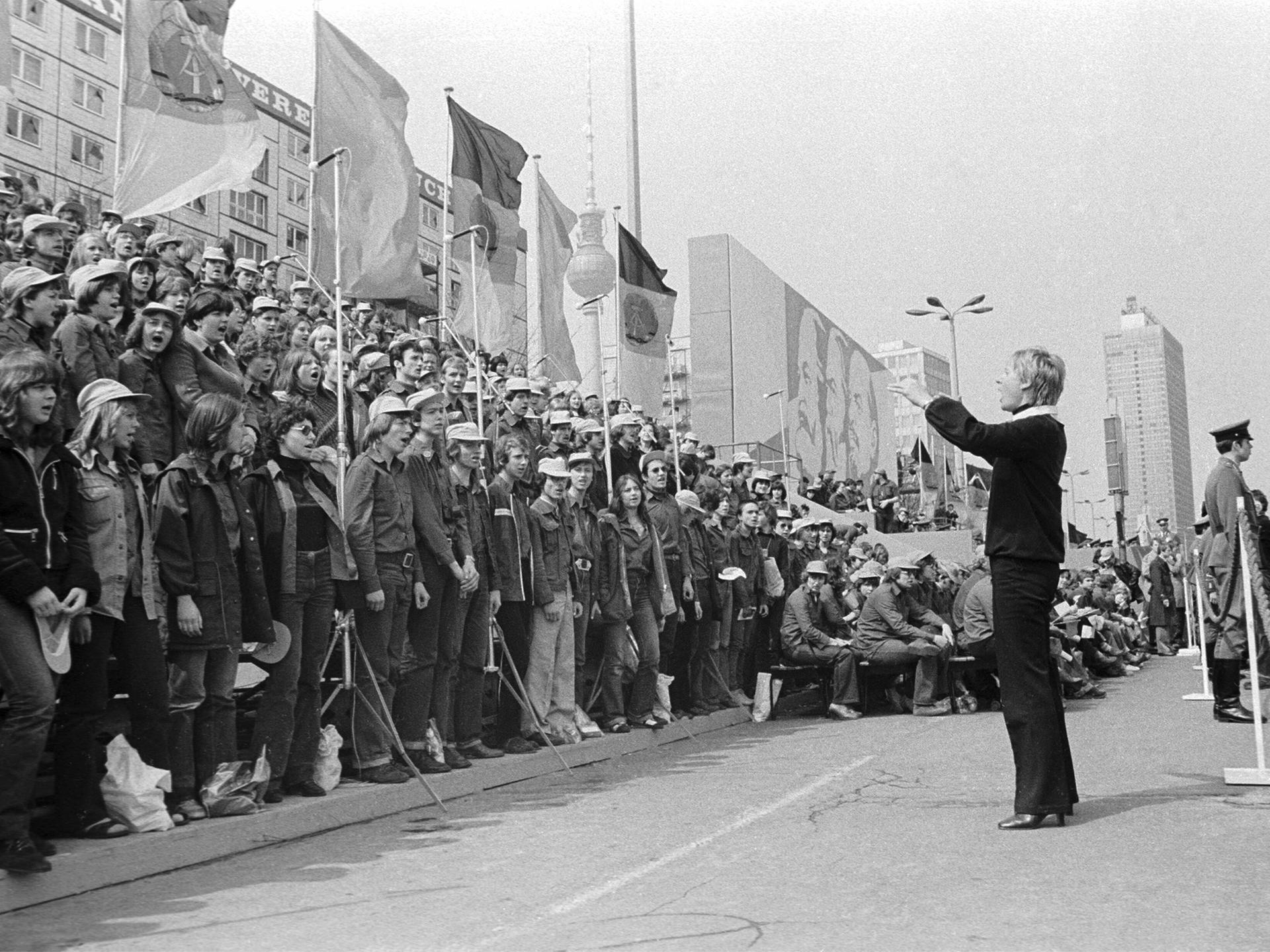 01.05.1980 DDR Ost-Berlin Stadtbezirk Mitte. Szene am Rande der Demonstration zum Ersten Mai. Eine junge Frau dirigiert während des Demonstrationszuges in der Karl-Marx-Allee einen Chor von Angehörigen der Freien Deutschen Jugend. , Berlin