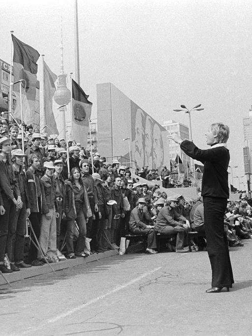 01.05.1980 DDR Ost-Berlin Stadtbezirk Mitte. Szene am Rande der Demonstration zum Ersten Mai. Eine junge Frau dirigiert während des Demonstrationszuges in der Karl-Marx-Allee einen Chor von Angehörigen der Freien Deutschen Jugend. , Berlin