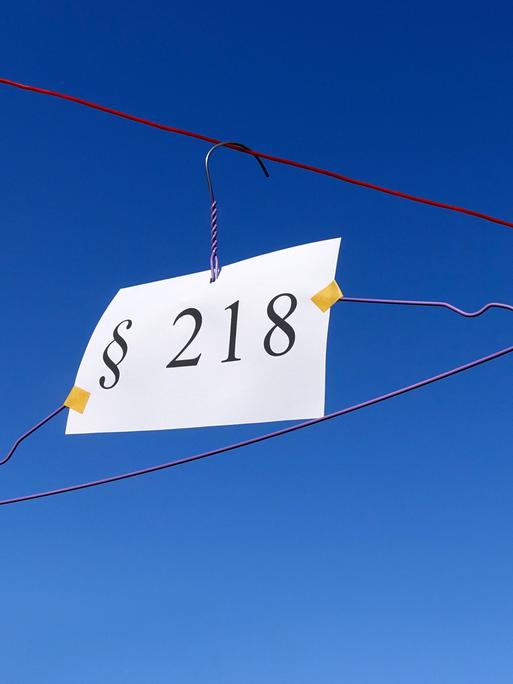 Vor blauem Himmel hängt ein Drahtbügel an einem Seil, daran hängt ein Schild auf dem Paragraf 218 steht.