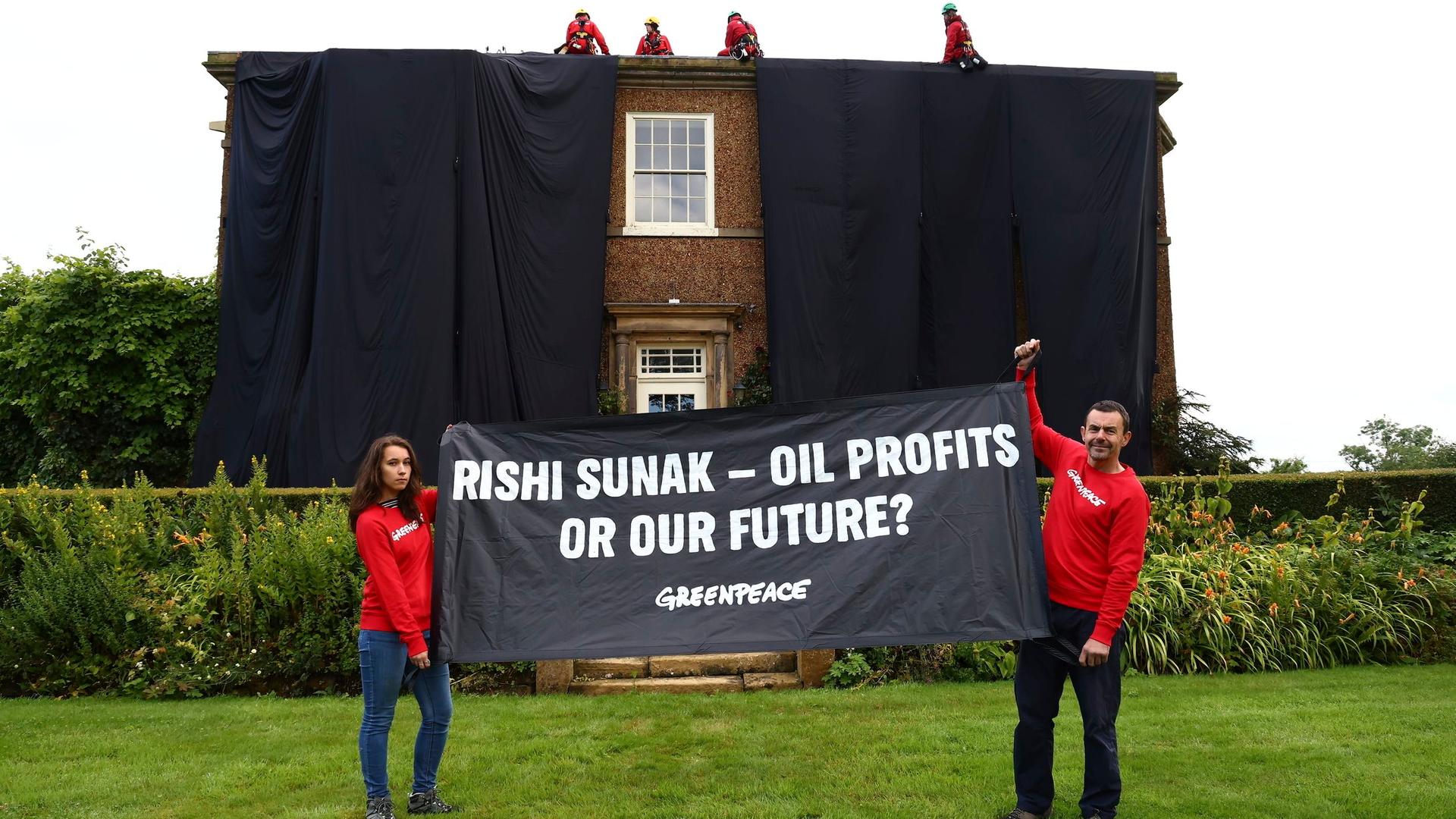 Das Haus des britischen Premierministers Sunak ist in schwarz verhüllt. Davor halten zwei Greenpeace-Aktivisten ein Transparent hoch, auf dem steht "Rishi-Sunak - Oil Profits or our Future?" Auf dem Dach sitzen vier weitere Aktivisten.