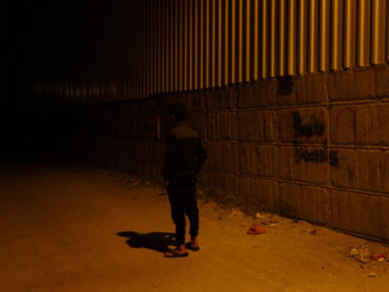 Ein schlanker schwarzhaariger Mann steht mit  dem Rücken zur Kamera vor einer hohen Mauer auf einer schwach beleuchteten Straße.