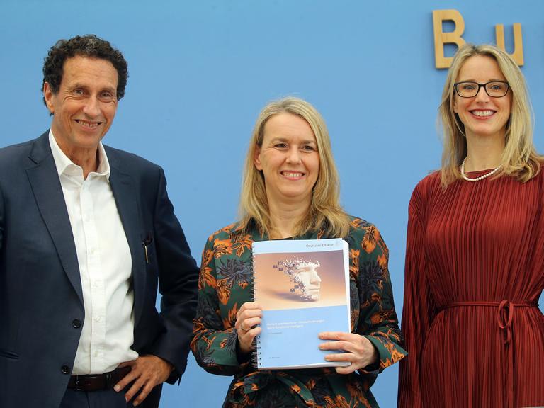 Das Foto zeigt 3 Forscher aus dem Ethik-Rat. Eine Forscherin hält ein Buch in die Höhe.