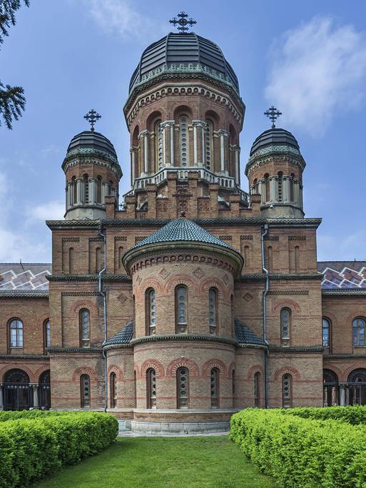 Ein Ziegelbau mit Türmen und Kuppeln steht in einem Park mit akkurat geschnittenen Hecken, die den Blick auf das Gebäude lenken.