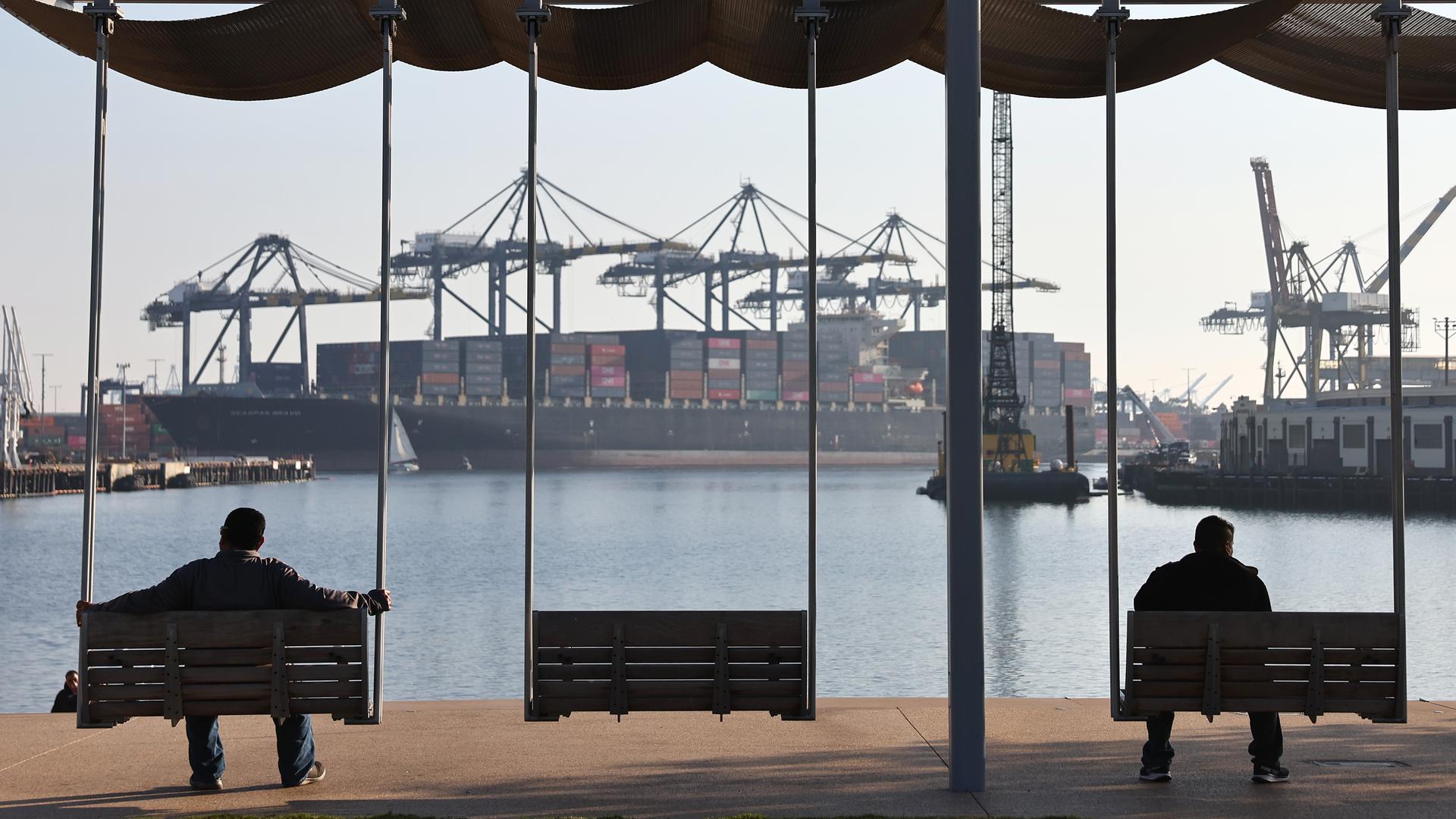 Zwei Personen sind als Silhouette zu sehen wie sie auf Bänken sitzend in Richtung eines Containerhafens mit einem großen Cargoschiff schauen.