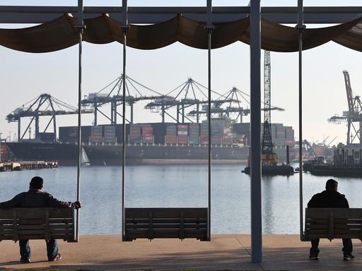 Zwei Personen sind als Silhouette zu sehen wie sie auf Bänken sitzend in Richtung eines Containerhafens mit einem großen Cargoschiff schauen.