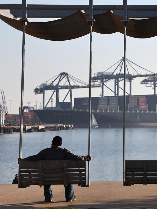 Zwei Personen sind als Silhouette zu sehen wie sie auf Bänken sitzend in Richtung eines Containerhafens mit einem großen Cargoschiff schauen.