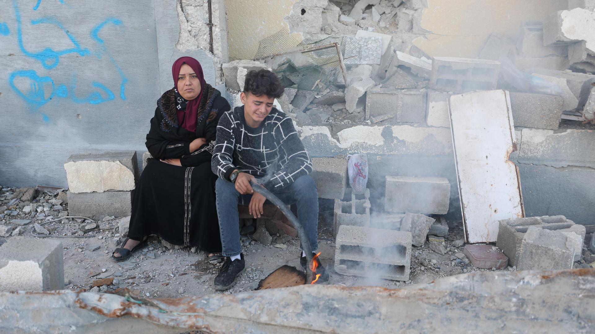 Eine Frau und ein Junge sitzen im Gazastreifen zwischen Trümmern, vor sich ein kleines Feuer.