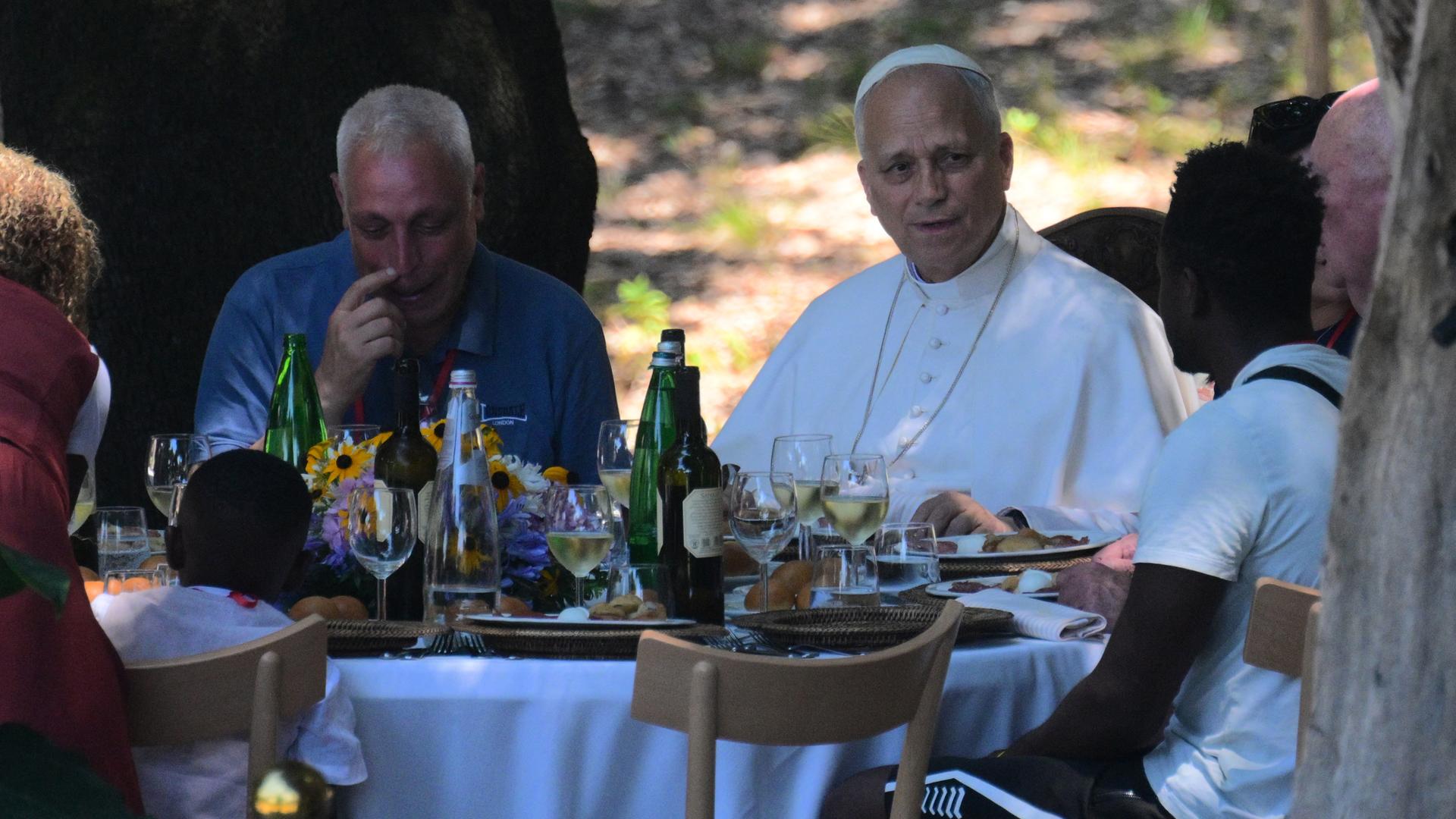 Der Papst sitzt mit mehreren weiteren Männern an einem Tisch. Auf dem Tisch stehen Speisen und Getränke. Der Papst spricht mit einem der Männer. Der Papst sitzt mit mehreren weiteren Männern an einem Tisch. Auf dem Tisch stehen Speisen und Getränke. Der Papst spricht mit einem der Männer.