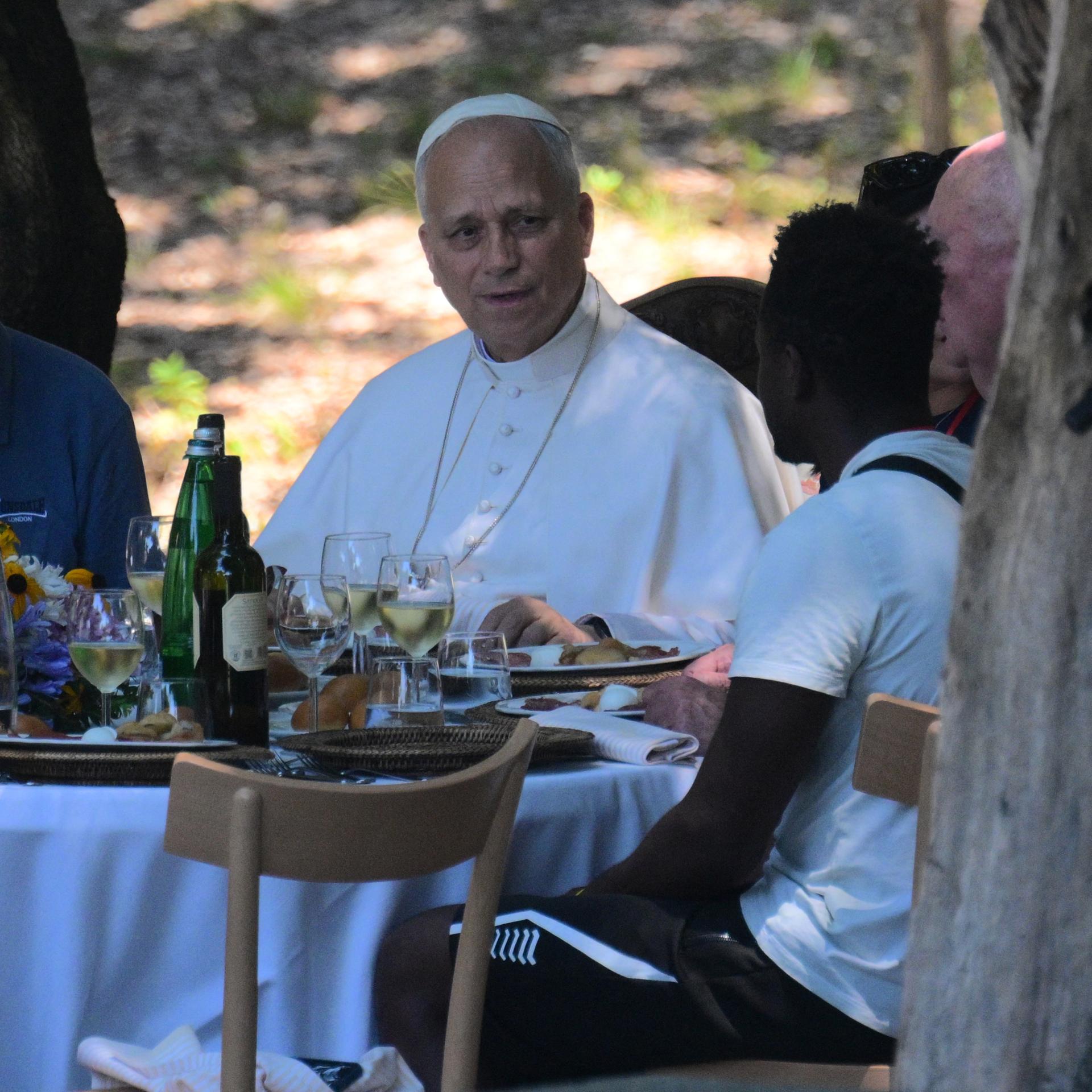 Der Papst sitzt mit mehreren weiteren Männern an einem Tisch. Auf dem Tisch stehen Speisen und Getränke. Der Papst spricht mit einem der Männer.