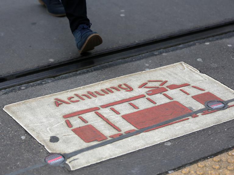 Das Licht einer Bodenampel leuchtet an einer Haltestelle, während ein Fußgänger die Straße kreuzt. Bodenampeln sollen Menschen, die gerade nach unten in ihr Smartphone blicken, vor Gefahren im Straßenverkehr warnen