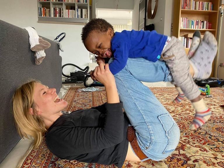 Ronja Buggel liegt auf dem Rücken und hält ihren Adoptivsohn Jeremiah auf den Knien, beide lachen. Der Junge hat Down-Syndrom. 