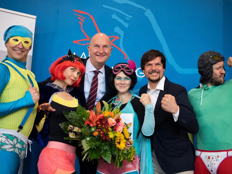 Trashig als Superhelden kostümierte Leute nehmen eine Auszeichnung entgegen und posieren dabei albern.