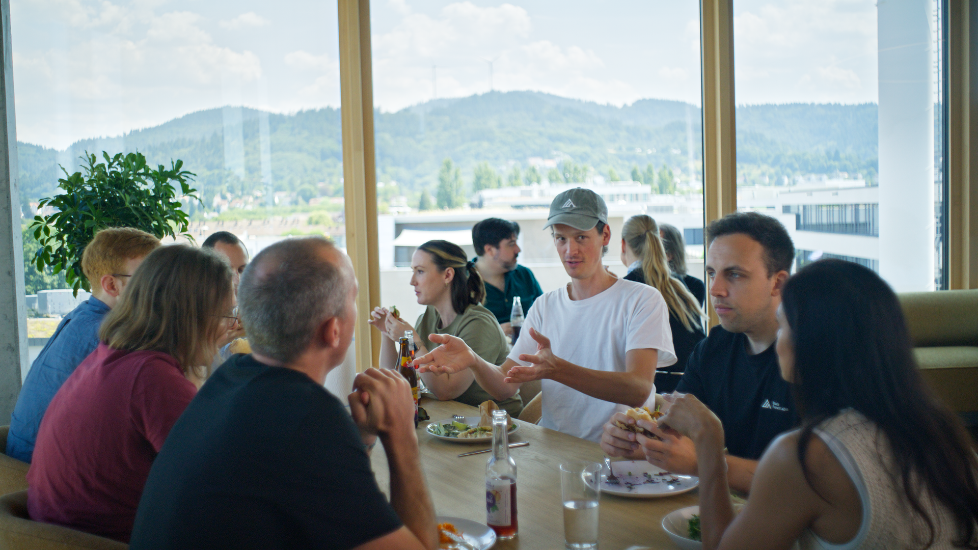 Mitarbeiter des Freiburger Startups Black Forest Labs sitzen an einem Esstisch in einer Kantine. Durch große Fenster sind im Hintergrund die Hügel des Schwarzwald zu sehen.