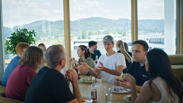 Mitarbeiter des Freiburger Startups Black Forest Labs sitzen an einem Esstisch in einer Kantine. Durch große Fenster sind im Hintergrund die Hügel des Schwarzwald zu sehen.
