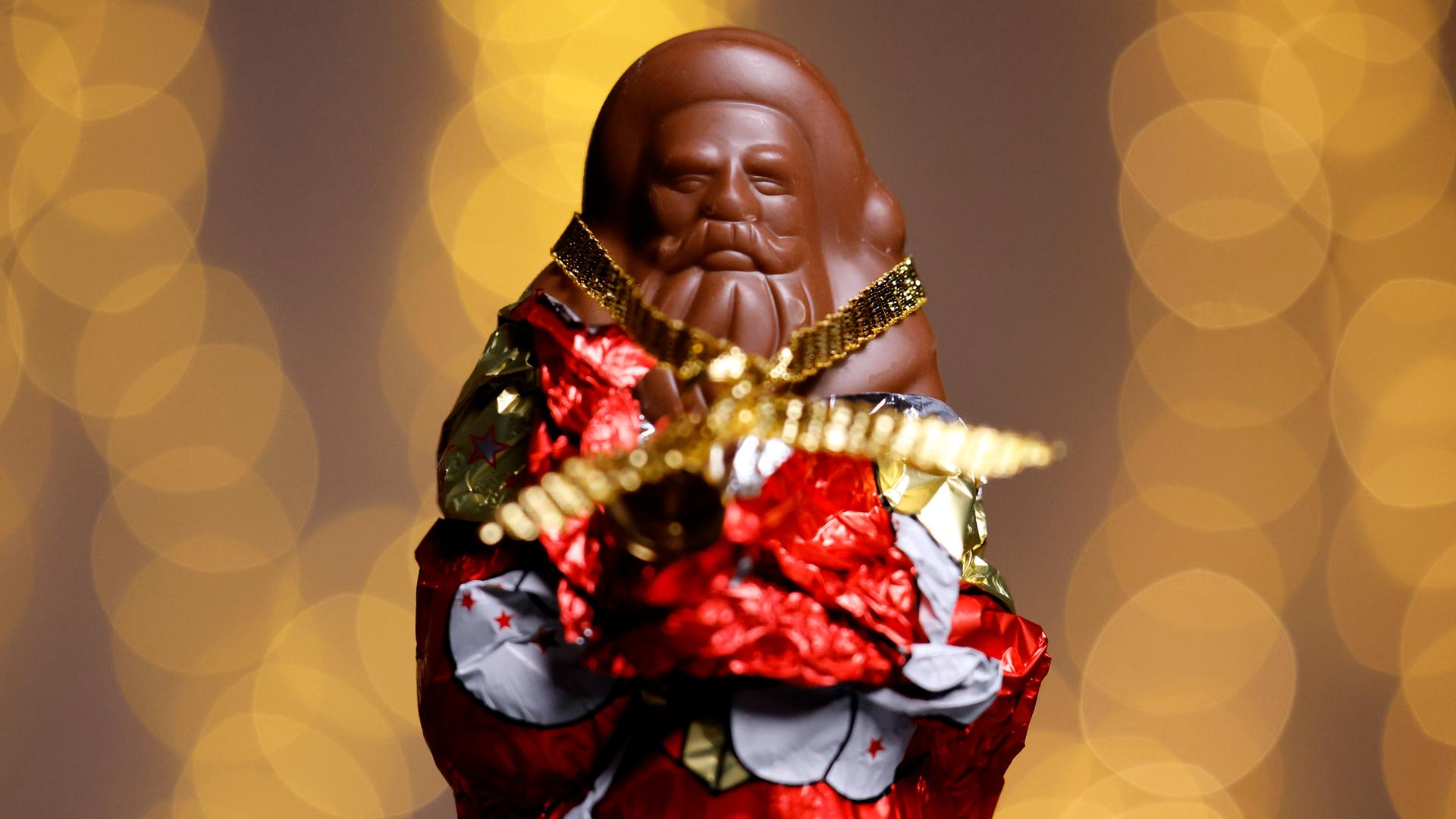 Ein teilweise ausgepackter Schokoladen-Weihnachtsmann in rot-goldener Folie. Der Hintergrund ist unscharf und besteht aus warmen, goldenen Lichtkreisen. (Symbolbild) Ein teilweise ausgepackter Schokoladen-Weihnachtsmann in rot-goldener Folie. Der Hintergrund ist unscharf und besteht aus warmen, goldenen Lichtkreisen. (Symbolbild)