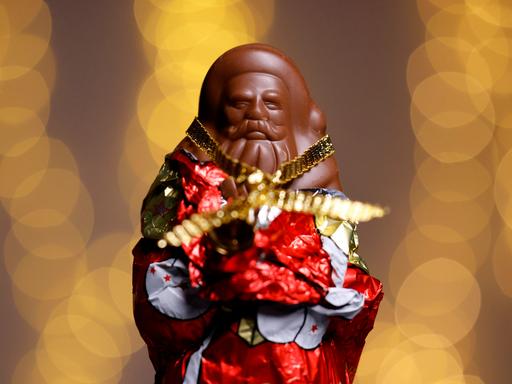 Ein teilweise ausgepackter Schokoladen-Weihnachtsmann in rot-goldener Folie. Der Hintergrund ist unscharf und besteht aus warmen, goldenen Lichtkreisen. (Symbolbild) 