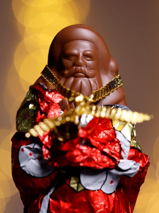 Ein teilweise ausgepackter Schokoladen-Weihnachtsmann in rot-goldener Folie. Der Hintergrund ist unscharf und besteht aus warmen, goldenen Lichtkreisen. (Symbolbild) 
