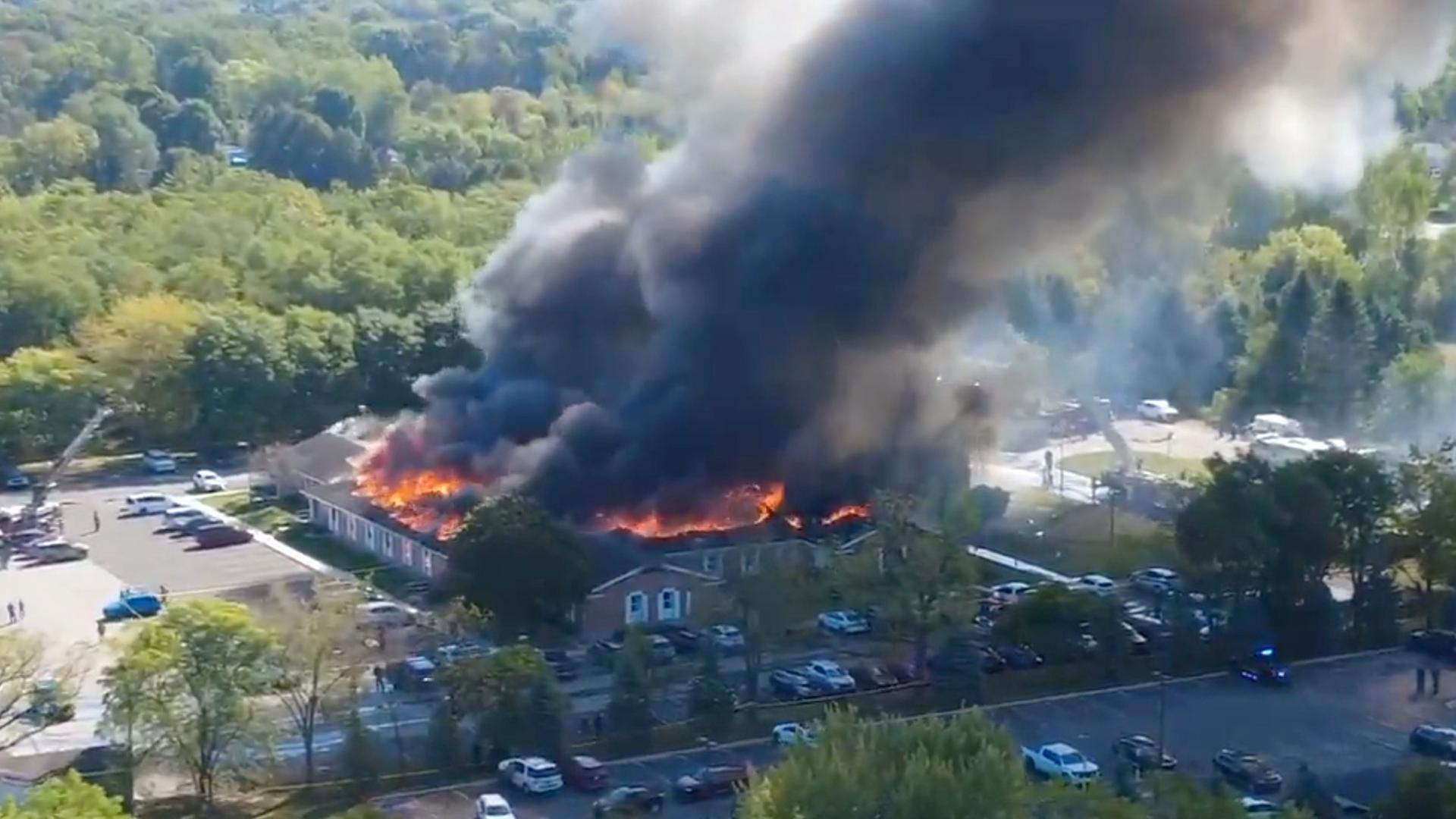 Blick aus der Hubschrauberperspektive: Das viereckigen Gebäude steht lichterloh in Flammen; schwarzer Rauch steigt aus. Davor viele geparkte Autos. 