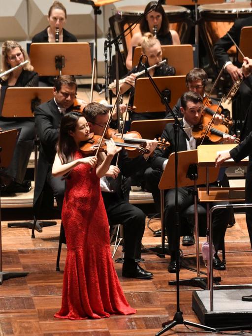 Geigerin Bomsori Kim trägt ein rotes Abendkleid und spielt ihre Geige. Im Hintergrund das Royal Philharmonic Orchestra. 