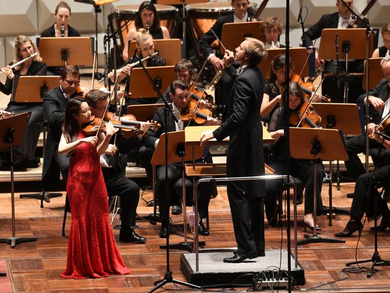 Geigerin Bomsori Kim trägt ein rotes Abendkleid und spielt ihre Geige. Im Hintergrund das Royal Philharmonic Orchestra. 