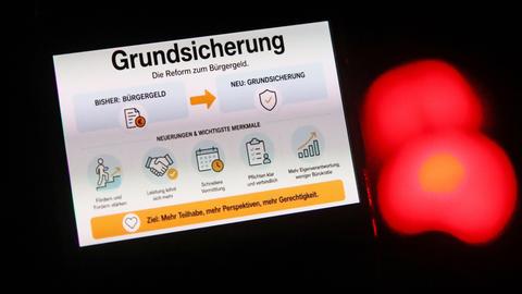 Ein Smartphone-Bildschirm zeigt eine Infografik zur Reform vom Bürgergeld hin zu einer neuen Grundsicherung, aufgenommen vor dem verschwommenen Hintergrund eines roten Lichts. 