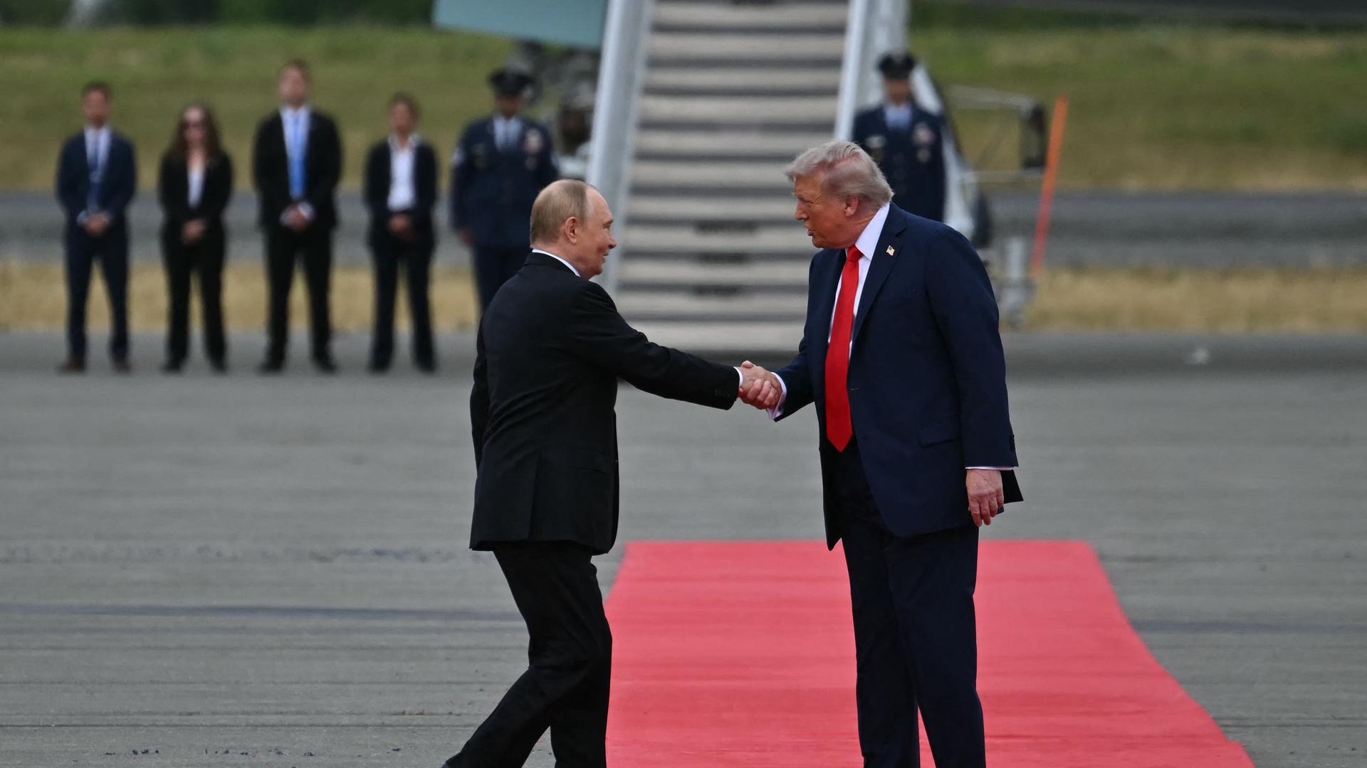 Putin wird von Trump begrüßt. Beide stehen auf einem roten Teppich und schütteln sich die Hand. 