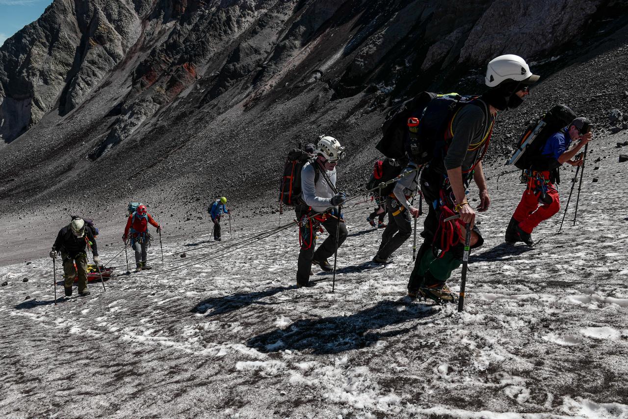 Menschen mit Bergsteigerausrüstung und Stöcken gehen einen schneebedeckten Hang hinauf.