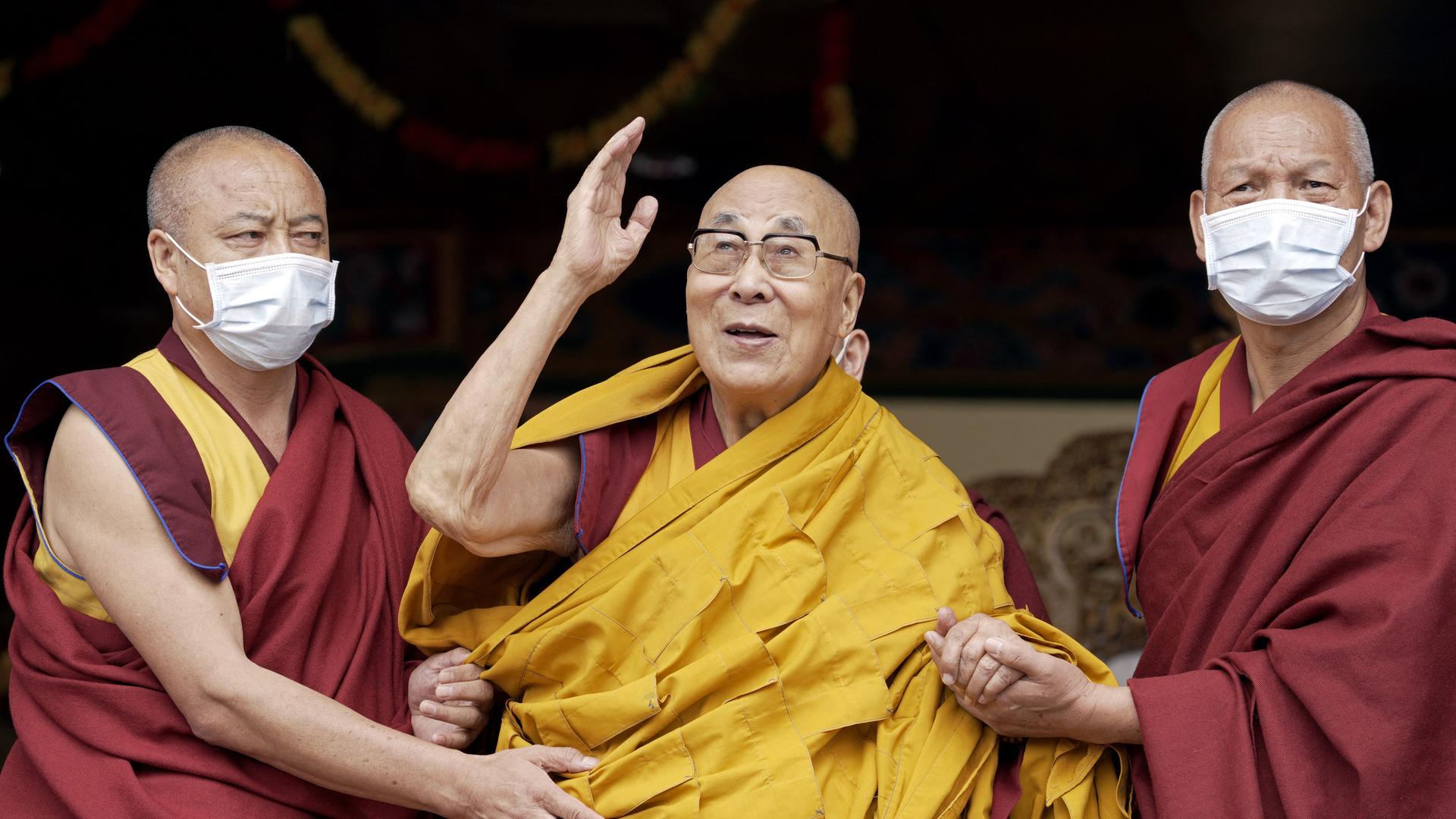 Der Dalai Lama winkt während einer Gebetszeremonie, während er von zwei Mönchen gestützt wird.