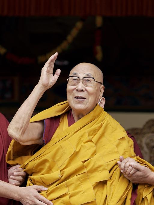 Der Dalai Lama winkt während einer Gebetszeremonie, während er von zwei Mönchen gestützt wird.