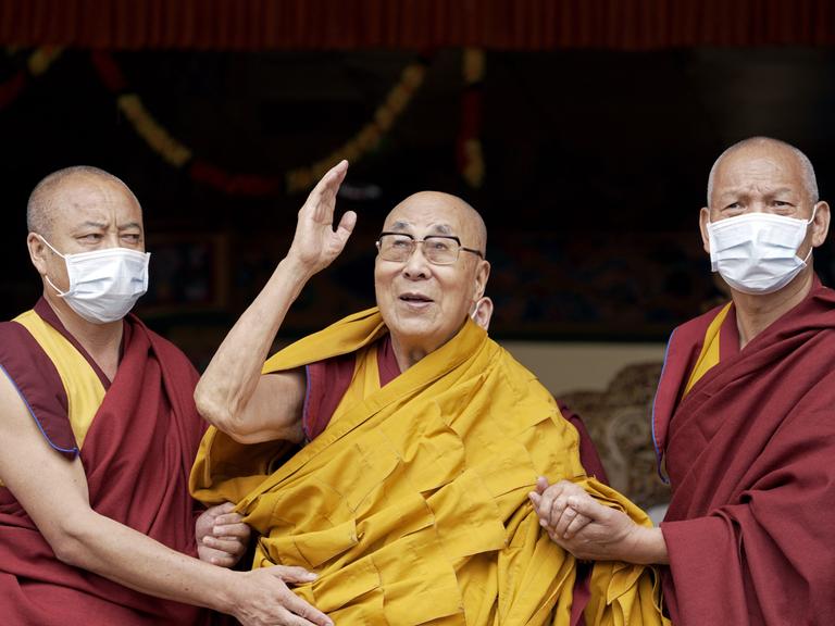 Der Dalai Lama winkt während einer Gebetszeremonie, während er von zwei Mönchen gestützt wird.