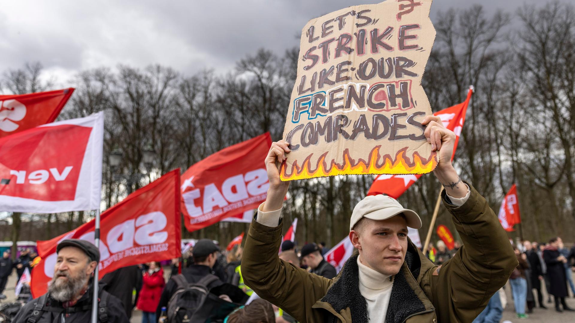 Eine ganze Reihe Personen nehmen an einem Gewerkschaftsprotest am 25. März 2023 teil. Ein Mann hält ein Schild hoch, auf dem steht "Let's strike like out French comrades".