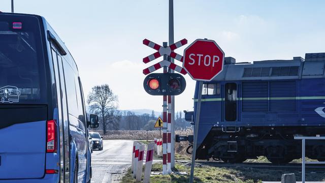 Ein Bahnübergang, dem sich ein Zug nähert. An dem Übergang blinkt ein rotes Licht. Auf der Straße halten ein Bus und ein Auto. 