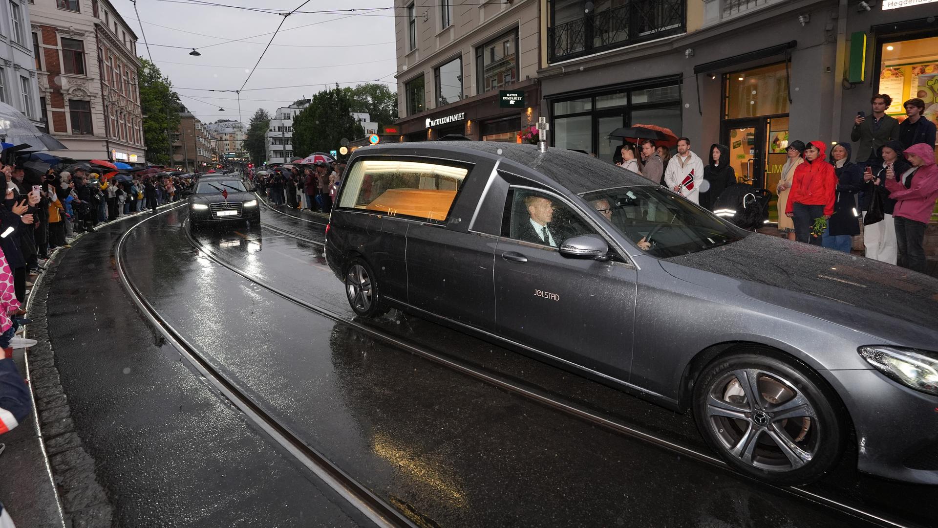 Auf dem Foto ist der Leichenwagen zu sehen, in dem sich der Sarg des verstorbenen Königs Harald V. befindet. Der Wagen fährt durch eine Einkaufsstraße; Menschen stehen am Rand der Straße und trauern. Auf dem Foto ist der Leichenwagen zu sehen, in dem sich der Sarg des verstorbenen Königs Harald V. befindet. Der Wagen fährt durch eine Einkaufsstraße; Menschen stehen am Rand der Straße und trauern.