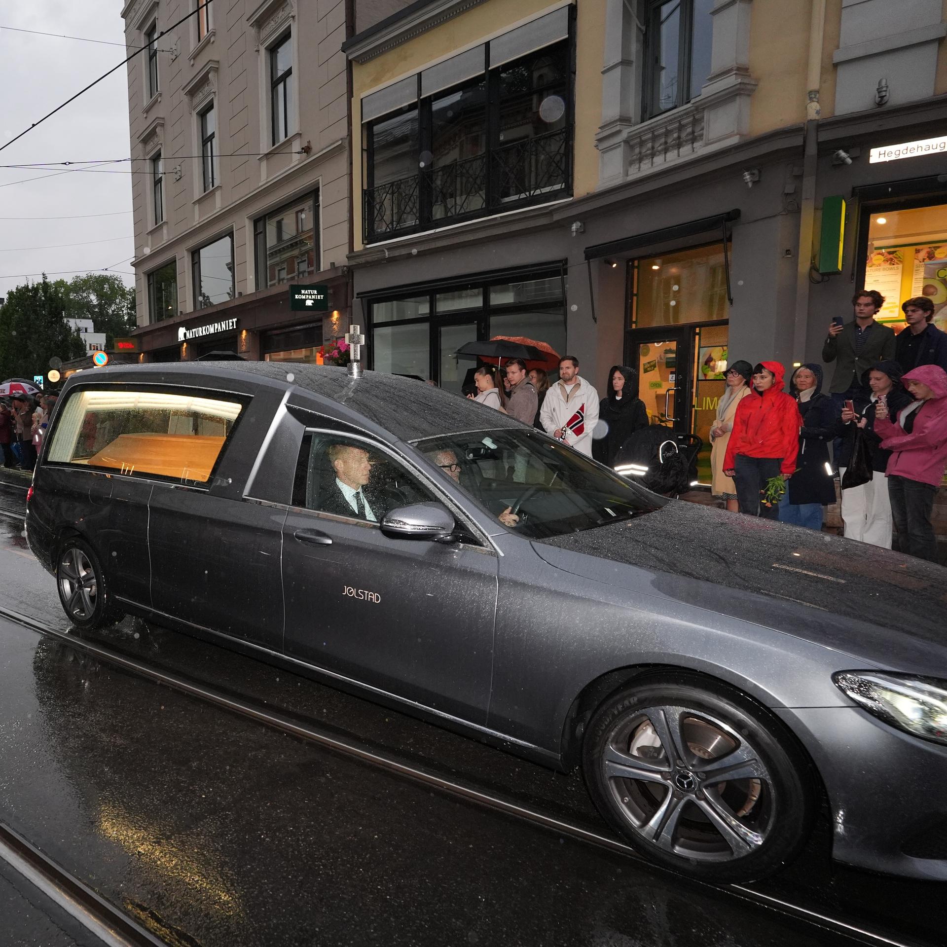 Auf dem Foto ist der Leichenwagen zu sehen, in dem sich der Sarg des verstorbenen Königs Harald V. befindet. Der Wagen fährt durch eine Einkaufsstraße; Menschen stehen am Rand der Straße und trauern.