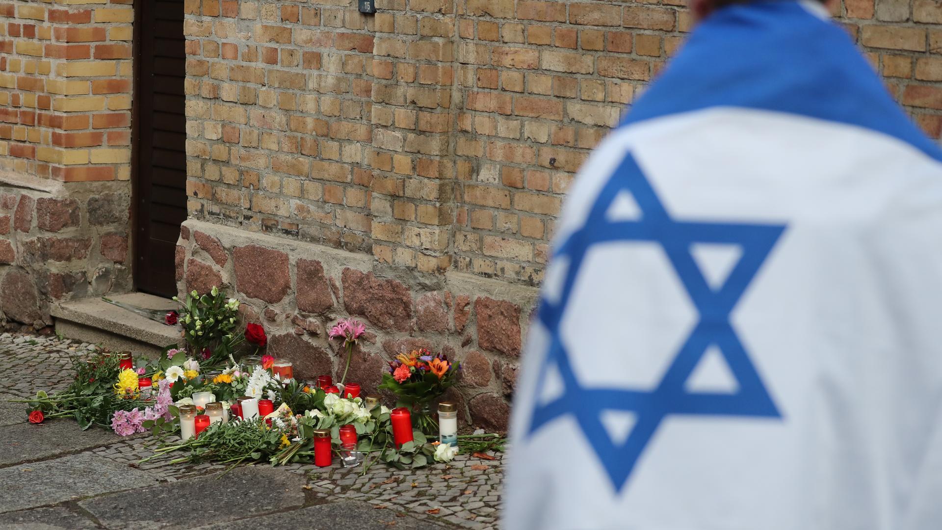 Ein Mann mit einer Israel-Flagge steht vor der Synagoge in Halle (Saale), wo Blumen und Kerzen stehen und liegen. 