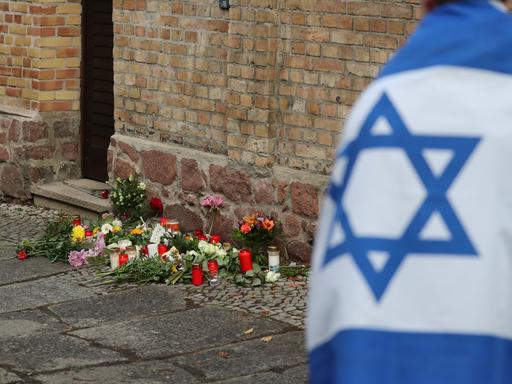 Ein Mann mit einer Israel-Flagge steht vor der Synagoge in Halle (Saale), wo Blumen und Kerzen stehen und liegen. 