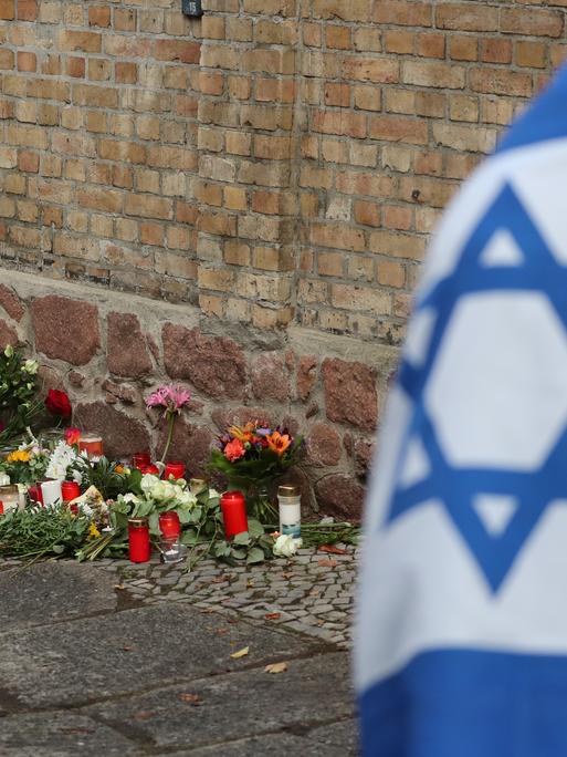 Ein Mann mit einer Israel-Flagge steht vor der Synagoge in Halle (Saale), wo Blumen und Kerzen stehen und liegen. 