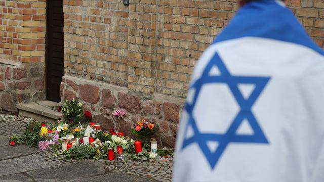 Ein Mann mit einer Israel-Flagge steht vor der Synagoge in Halle (Saale), wo Blumen und Kerzen stehen und liegen. 