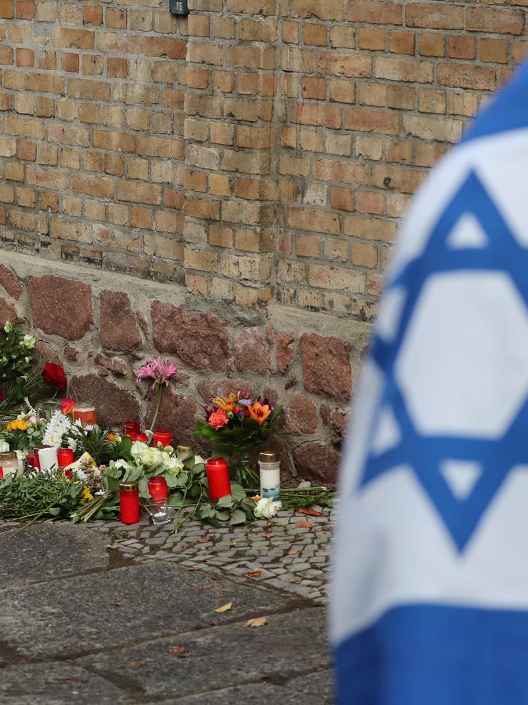 Ein Mann mit einer Israel-Flagge steht vor der Synagoge in Halle (Saale), wo Blumen und Kerzen stehen und liegen. 