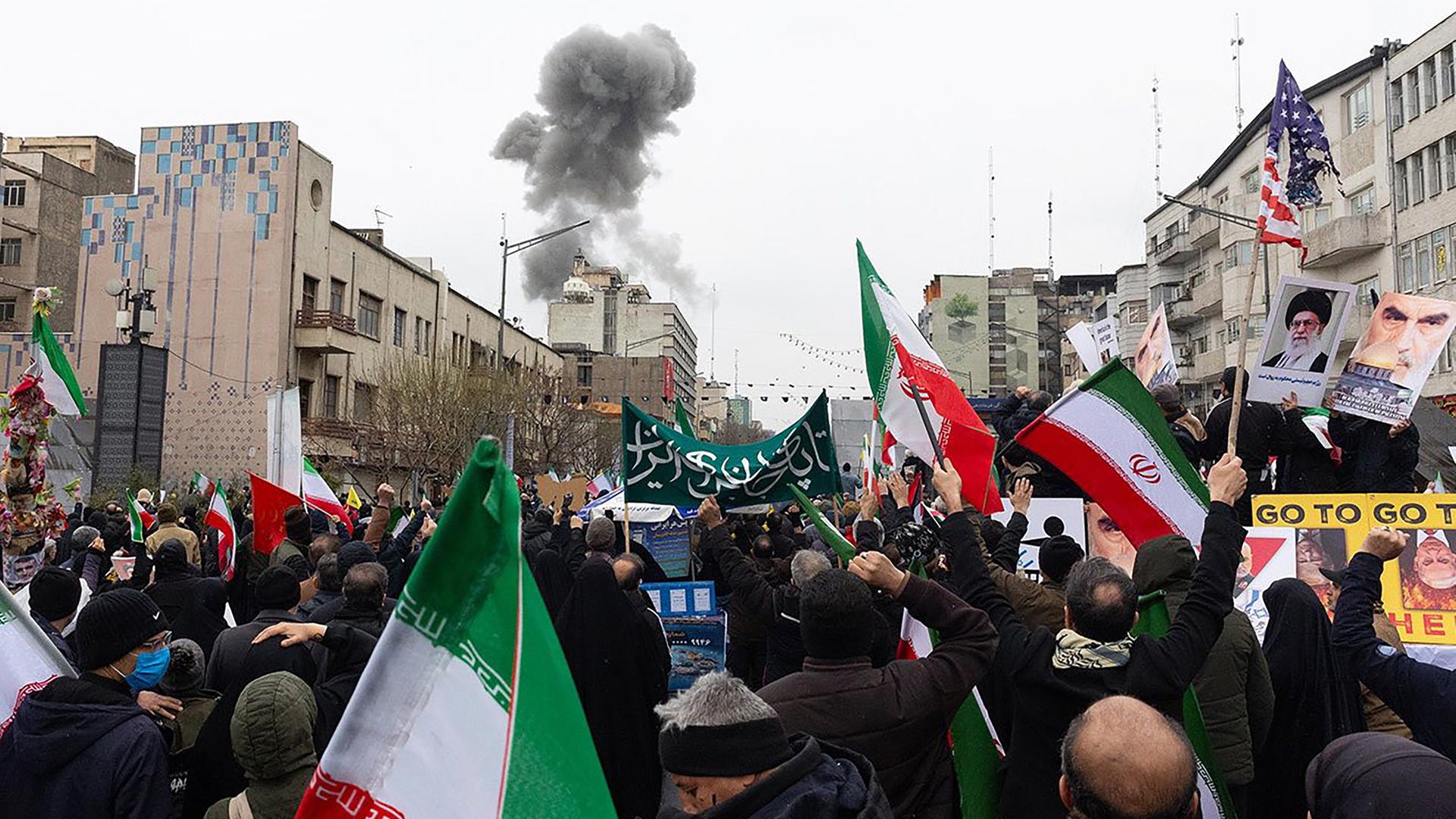Viele Menschen bei einer Kundgebung, im Hintergrund steigt eine Rauchsäule auf.
