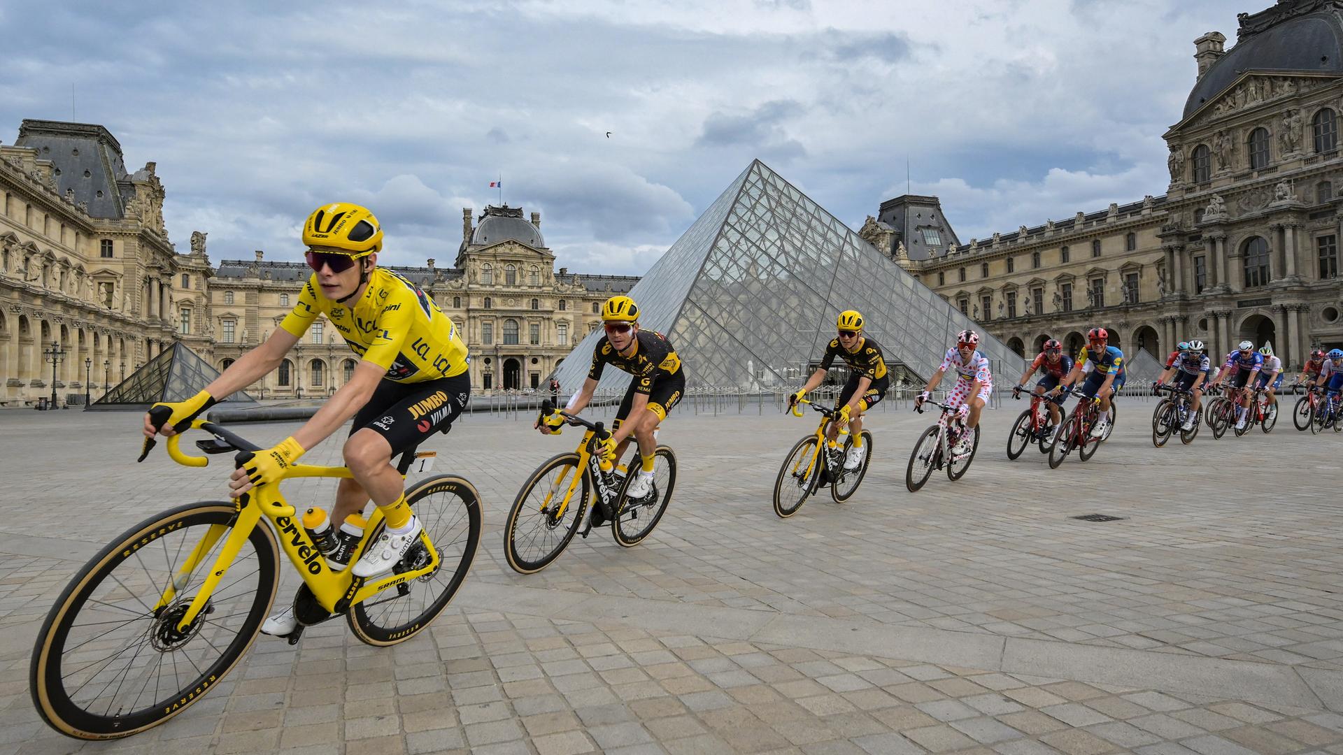 Tour de France-Sieger Jonas Vingegaard im gelben Trikot vor dem Louvre in Paris. Hinter ihm folgen einige andere Radfahrer.